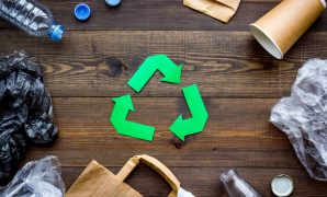Green paper recycling sign among waste materials paper, plastic, polyethylene on dark wooden background top view.